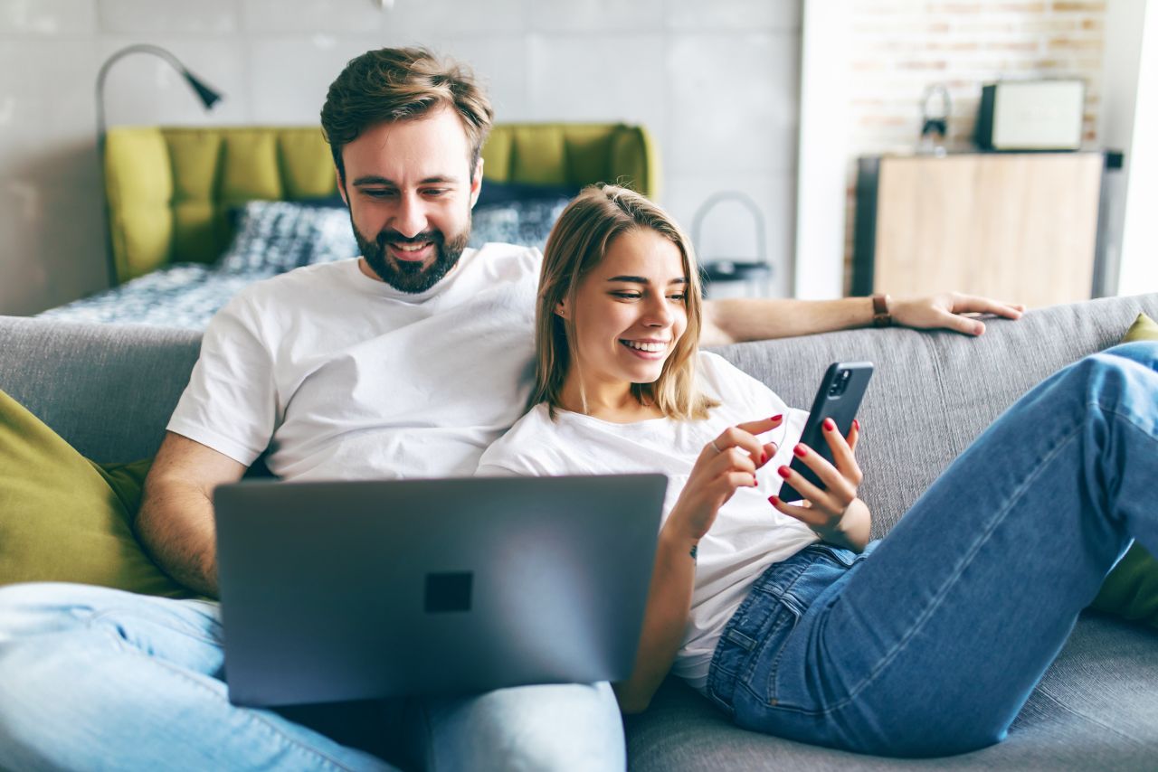 Beautiful young couple relaxing on a couch at home using laptop computer and mobile phone