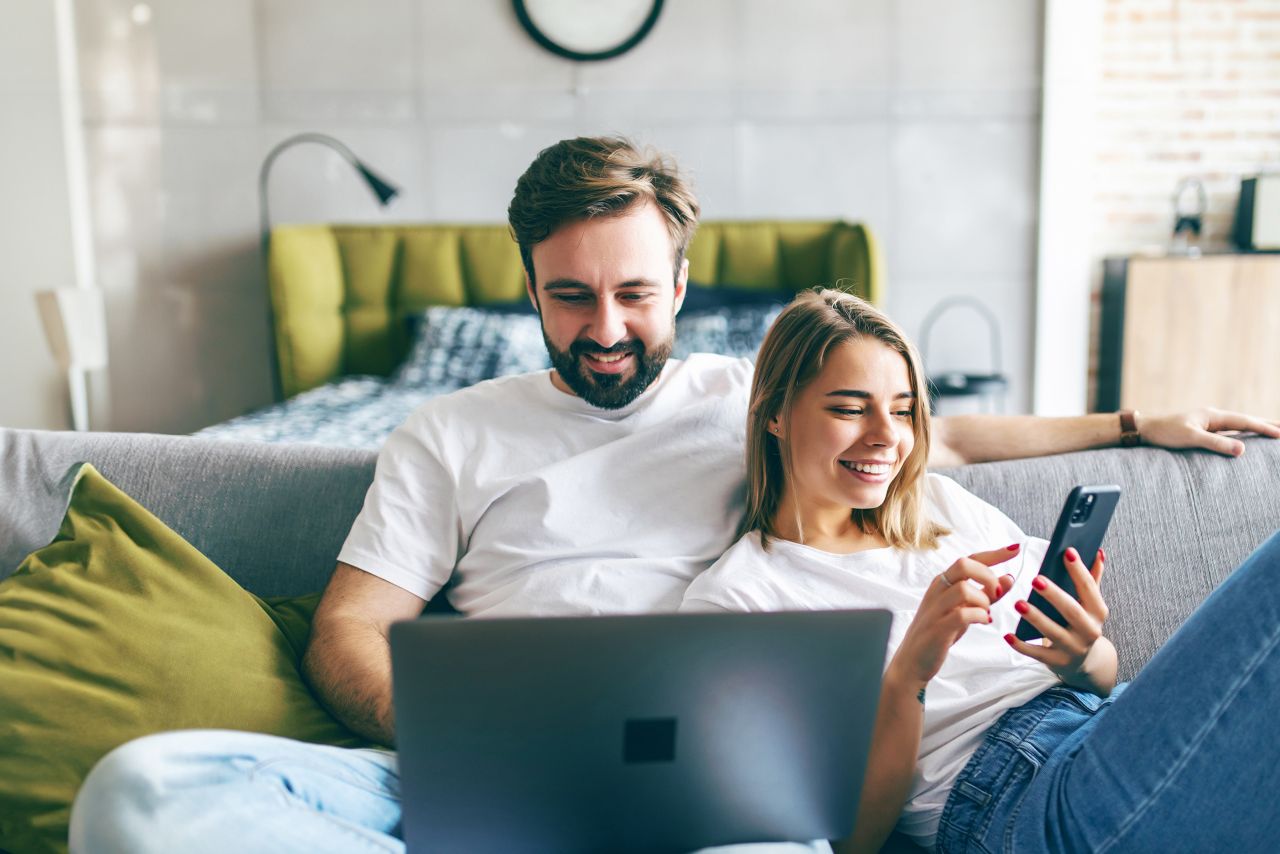 Beautiful young couple relaxing on a couch at home using laptop computer and mobile phone